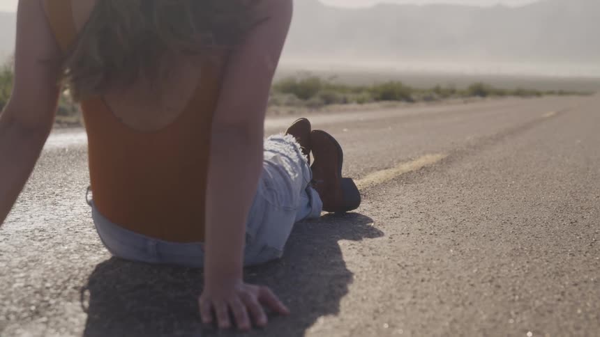 Young woman sits on rural highway through desert and mountains