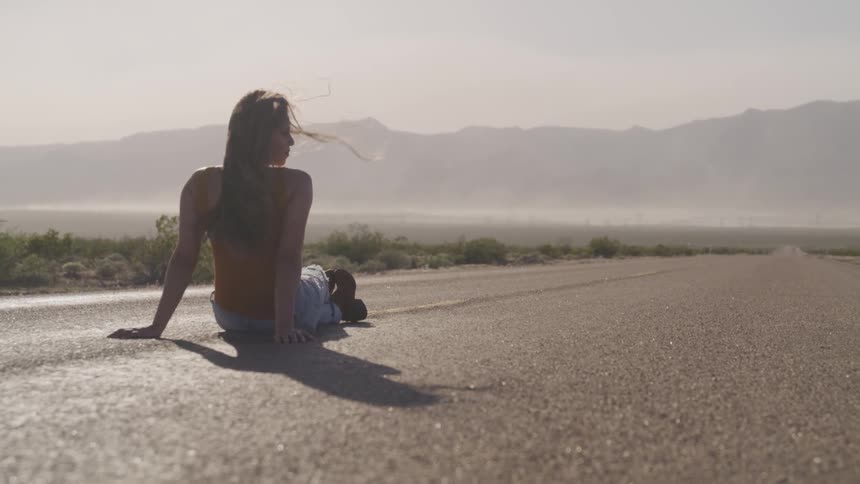 Young woman sits on empty rural highway in front of mountains