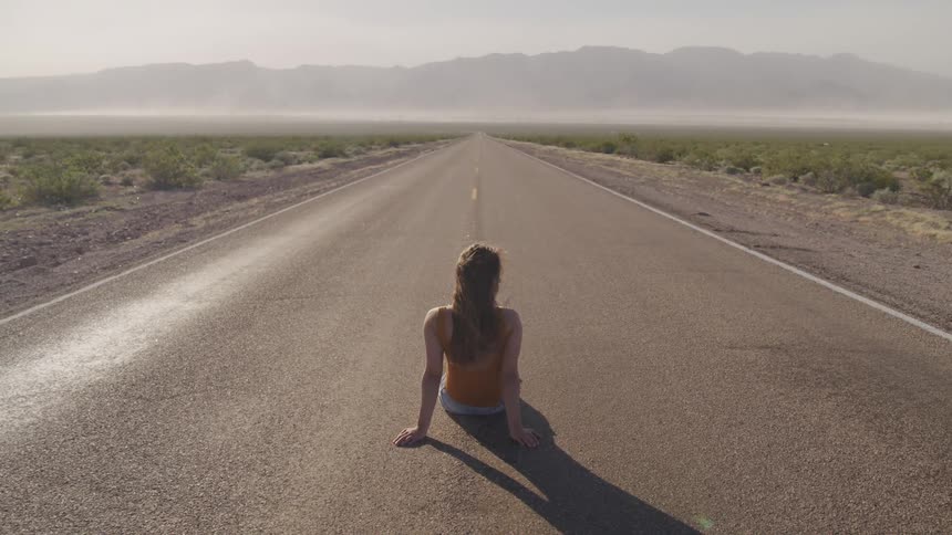 A young woman sitting in the middle of a quiet road with fields on either side
