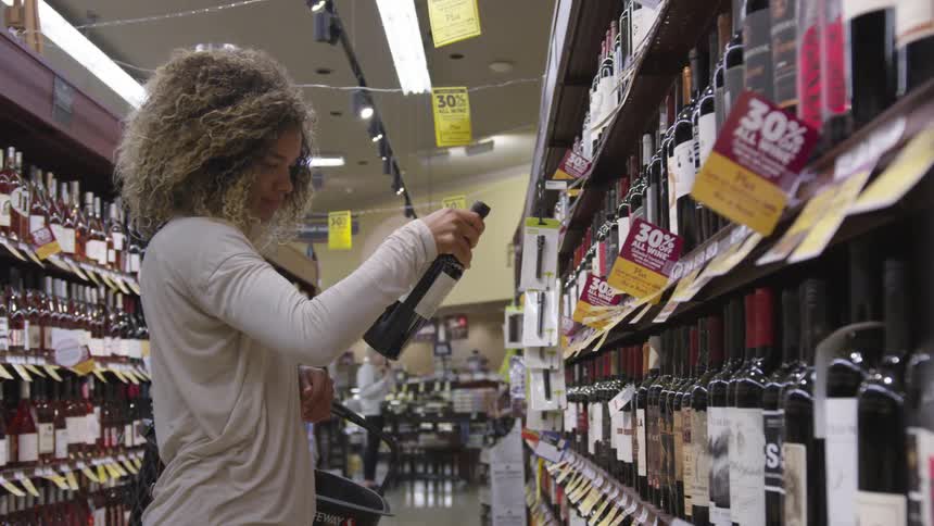 A woman standing in a store aisle carrying a basket reaches and pulls a wine bottle off a shelf