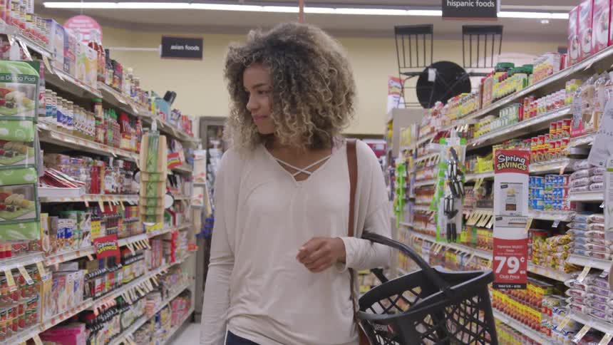 A young woman carries a shopping basket on her arm as she examines the shelves in an aisle of a grocery store