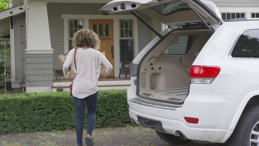A woman carries groceries from the back of vehicle towards a house