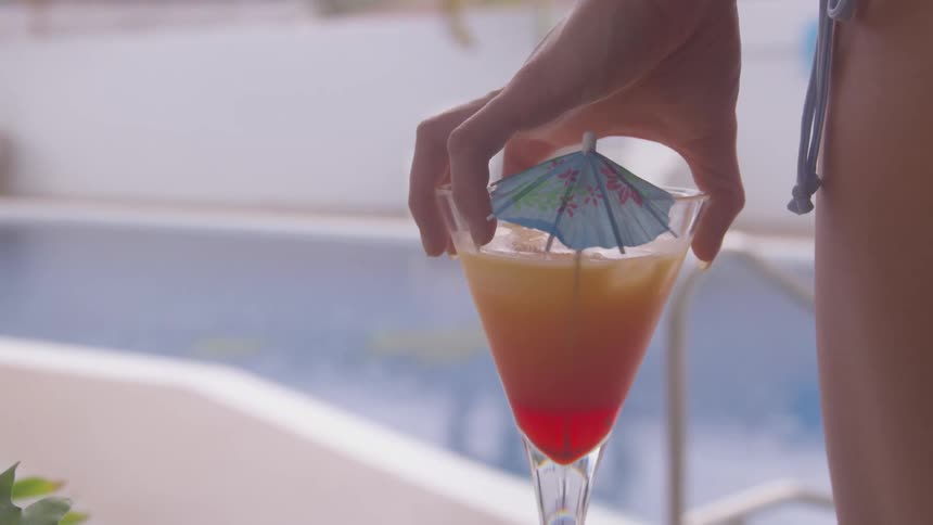 A young woman stands by an outdoor pool holding a cocktail