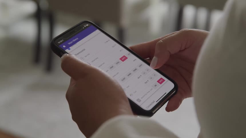 A woman adjust settings on a thermostat app on a smartphone by tapping the screen with her thumb