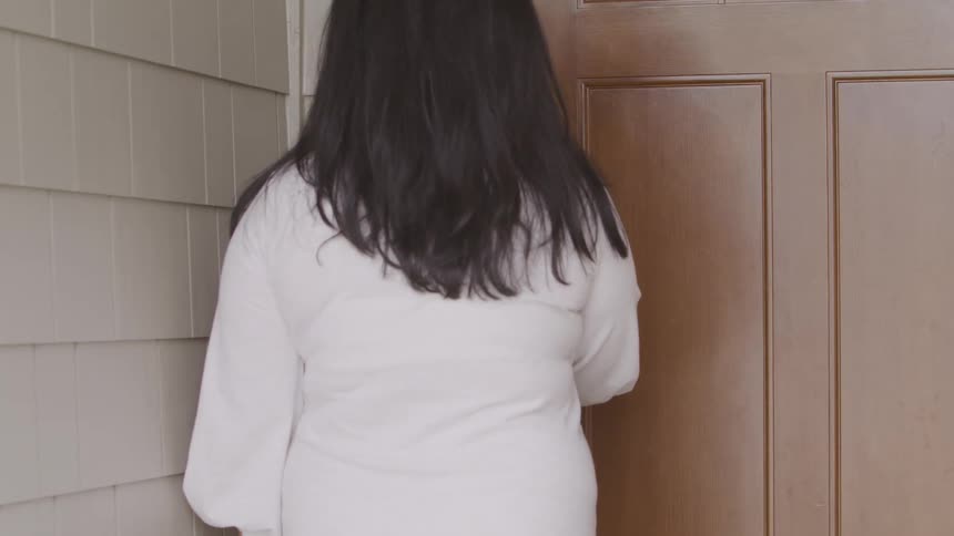 An asian woman in a bath robe enters a code on a keypad to unlock the front door of a house before entering it