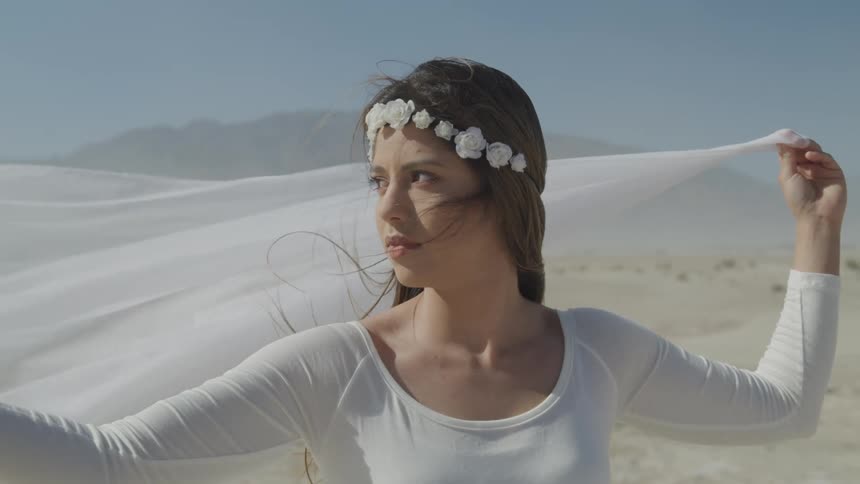 A teenage girl stands on a beach and holds a scarf behind her head as it blows in the wind