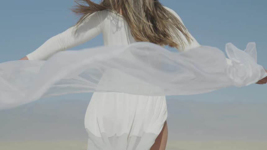 A woman stands on the beach and holds her scarf behind her back as the wind blows it