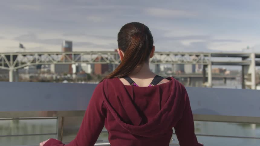 Woman stands at rail looking across water toward bridge and city
