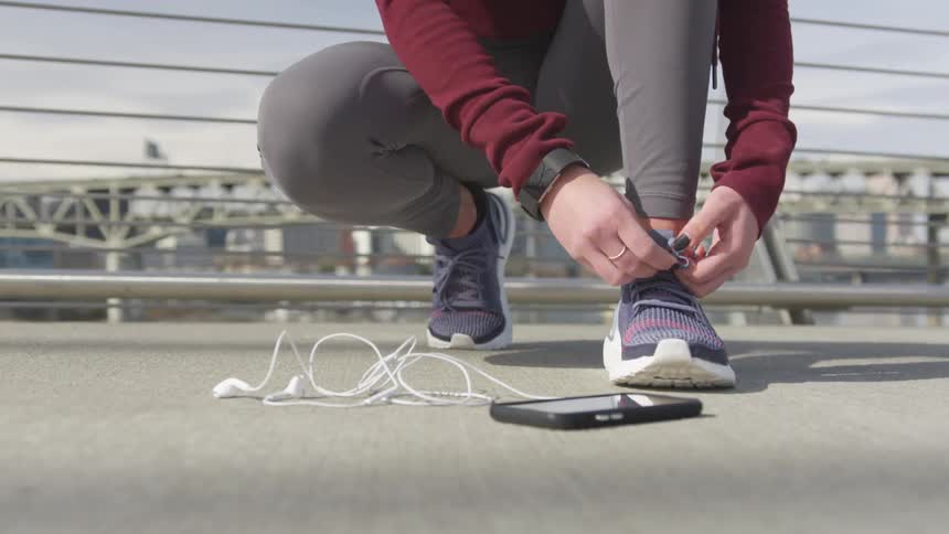 A woman in athletic wear kneels and ties her shoes while on a bridge on a sunny day