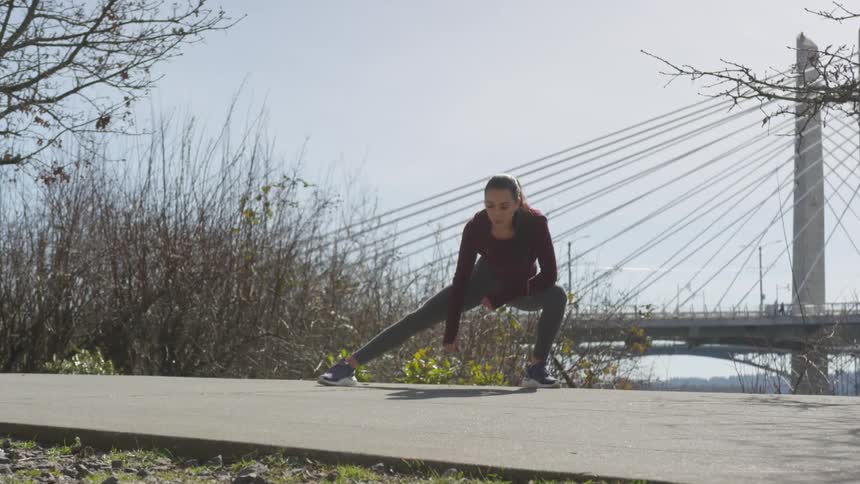 A woman rocks back and forth stretching her legs on a pedestrian pathway with a large bridge in the background