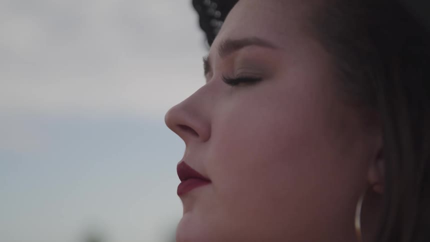 A woman in red lipstick stands outdoors during the day with her eyes closed