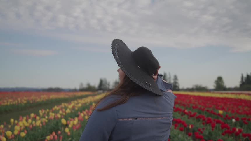 A woman with a hat standing in front of a flower field with trees and the sky in the background