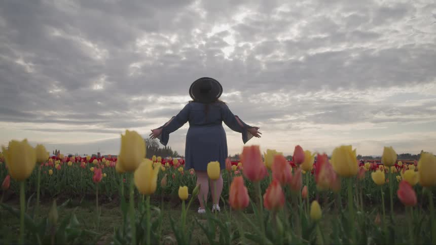 A woman stands in field of pink and yellow flowers and faces the cloudy sky with her arms held wide