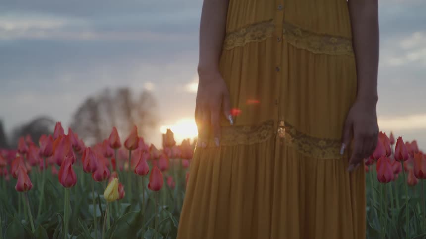 A woman in a yellow dress standing in a flower field with trees the sky and clouds in the background