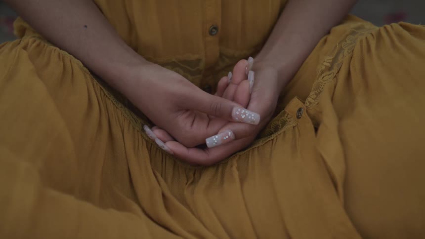 Woman wearing dress with long manicured nails sits cross legged with hands clasped in her lap
