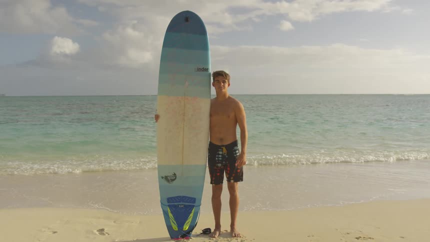 A teenage male surfer stands and poses on the beach with a colorful long surfboard