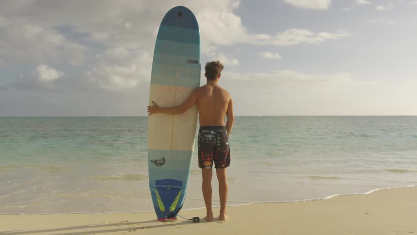 Male holding up surf board standing on shoreline looking out to horizon