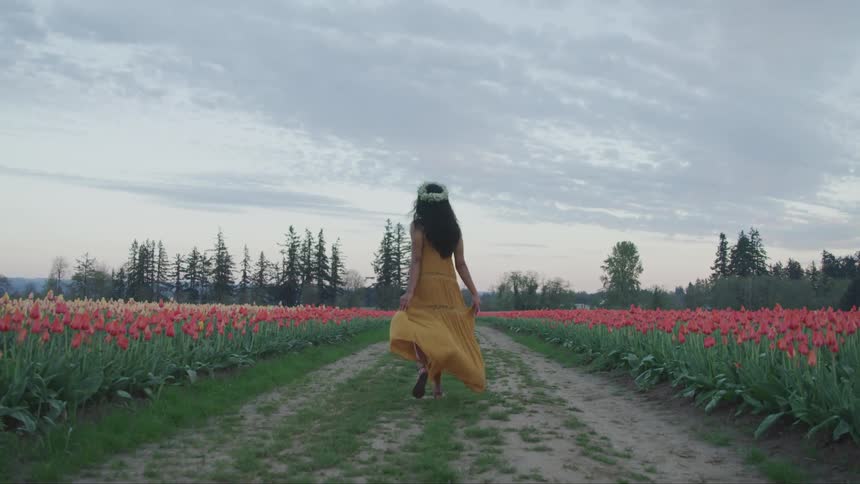 A young woman spins and skips along a dirt path through a field while holding the helm of her dress during the day