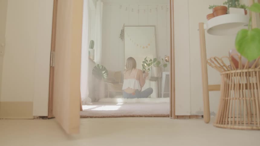 A woman applies makeup to her face while sitting on the floor in front of a full length mirror in her house