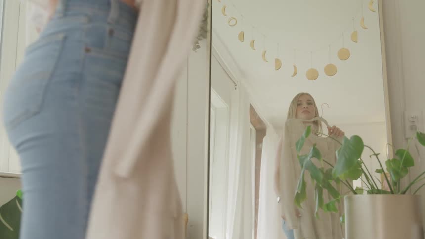 A young woman holds a shirt in front of herself as she poses in a mirror