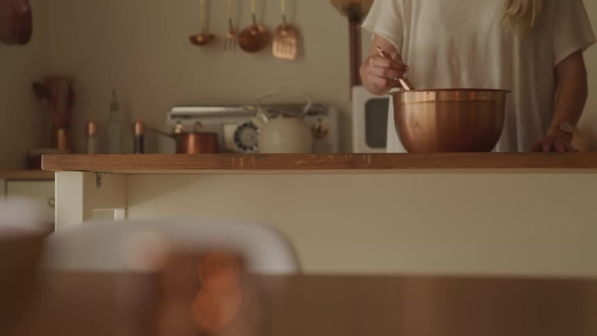 A woman whisks the contents of a bronze bowl in a kitchen full of bronze cooking ware