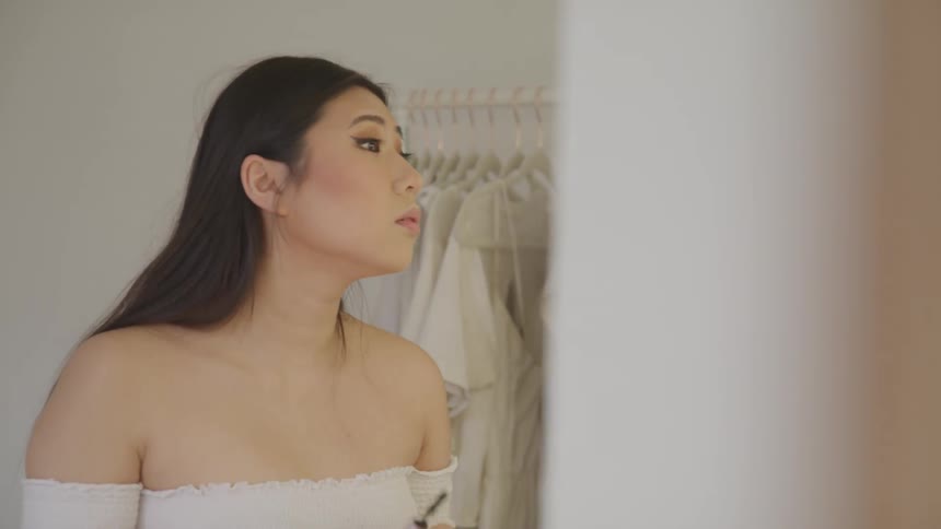 A woman standing in front of a mirror applying makeup with clothing in the background