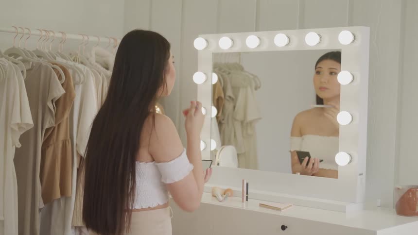 Young woman applies makeup in front of dressing room mirror with lights