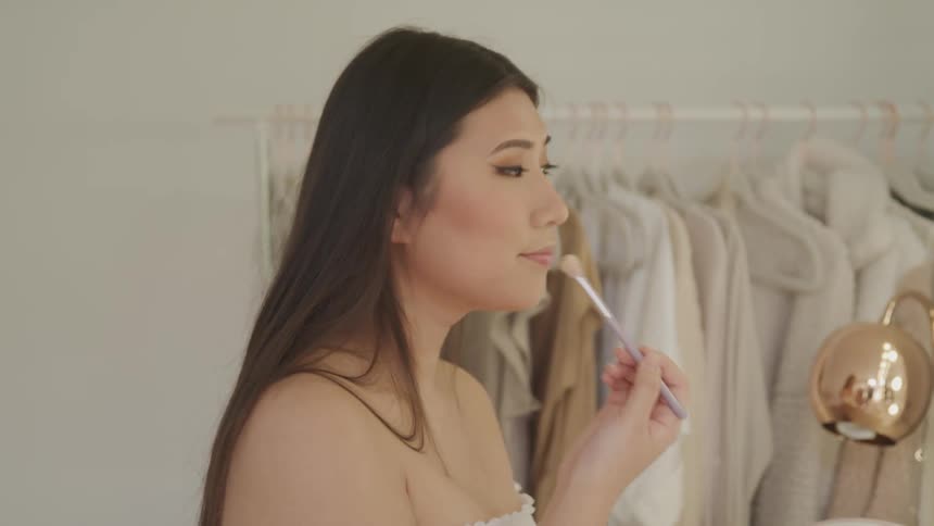 A woman applies power a brush in an all white dressing room with a lighted vanity