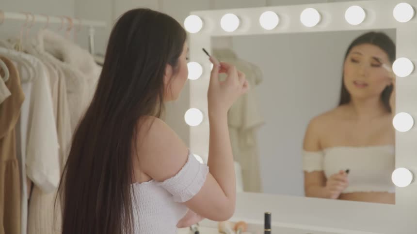 A young asian woman applies mascara while looking into the mirror of her vanity