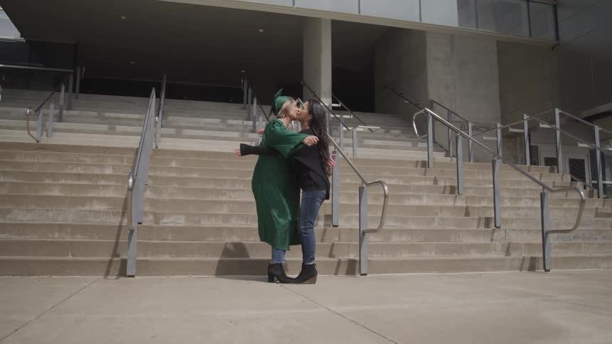 A young woman in a graduate gown and cap embraces her mother outside of a large building during the day