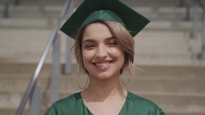 A young woman in a graduate gown and cap smiles into the camera while standing outdoors in front of a cement staircase