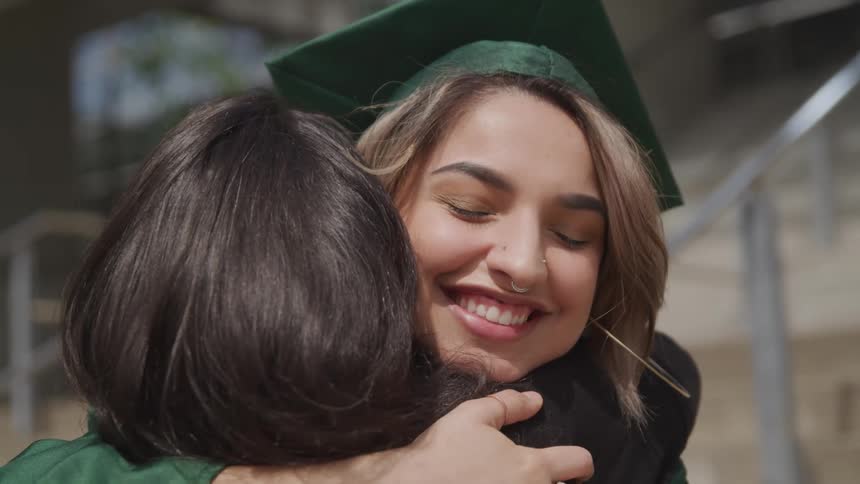A young woman in a graduate gown hugs another woman at the bottom of a cement staircase outdoors during the day