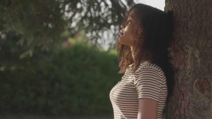 A young woman looks up at the sky while resting her back against a tree on a sunny day