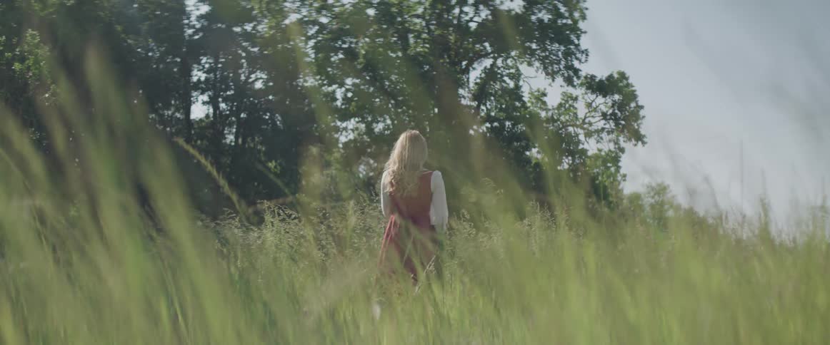 A woman in a dress stands alone in a tall grassy field lined with trees in the daytime