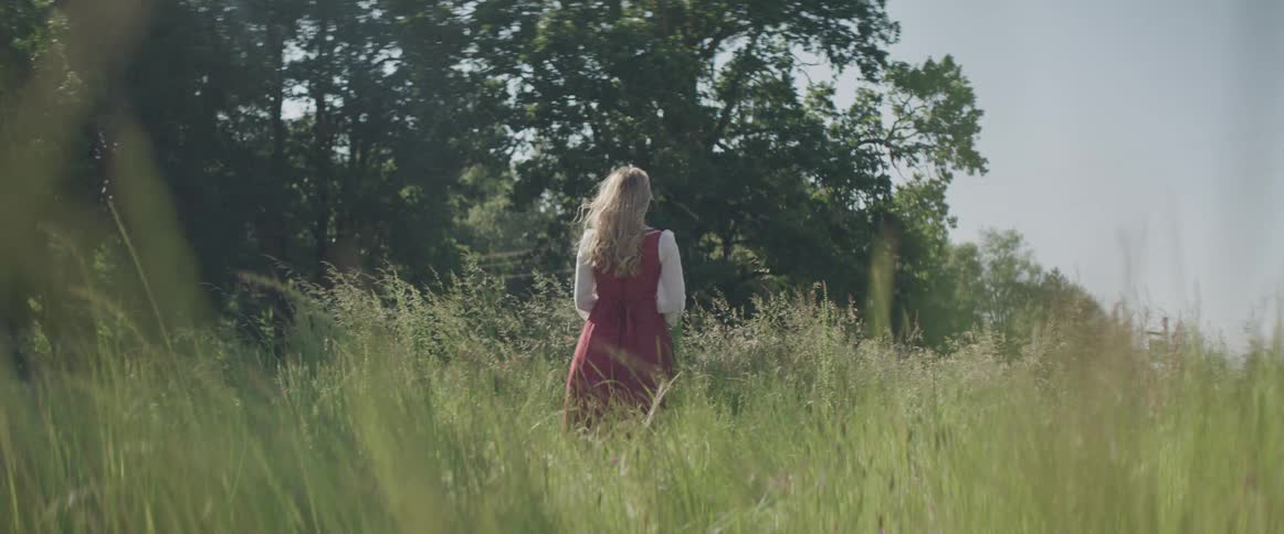 A woman in a dress standing in a grassy field with trees and the sky in the background
