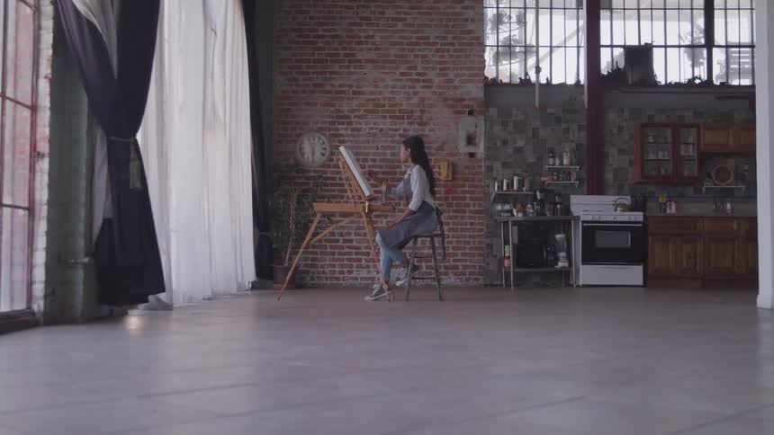 A female artist painting on a canvas in an art studio while sitting on a stool