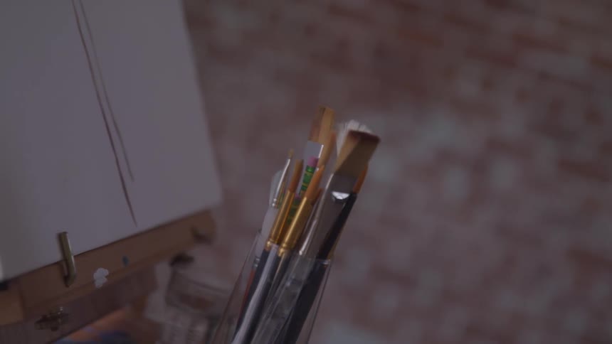 A group of clean paint brushes stand together in a container next to a canvas