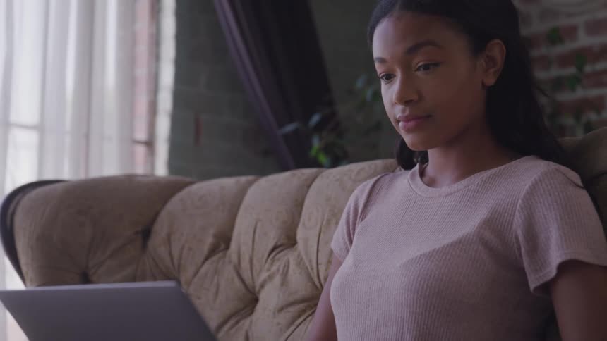 A woman sitting on a couch typing on a laptop with a wall in the background