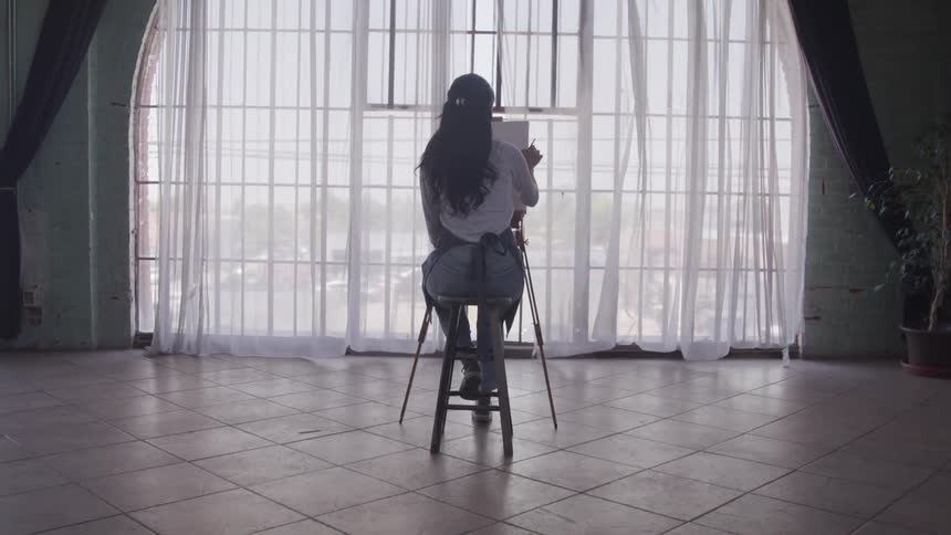 A woman sits on a stool painting on an easel facing a large window