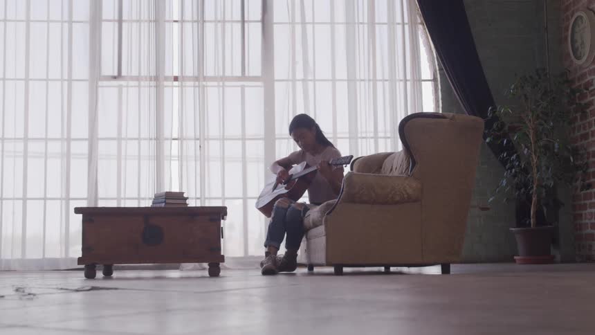 A young woman plays a guitar while sitting on a sofa in front of a large window in the living room of a house