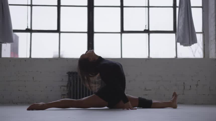 A woman doing a split in a room with a window and curtains in the background