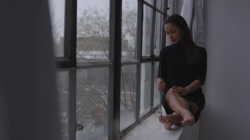 A barefoot woman sits on the sill of a large window while lost in thought