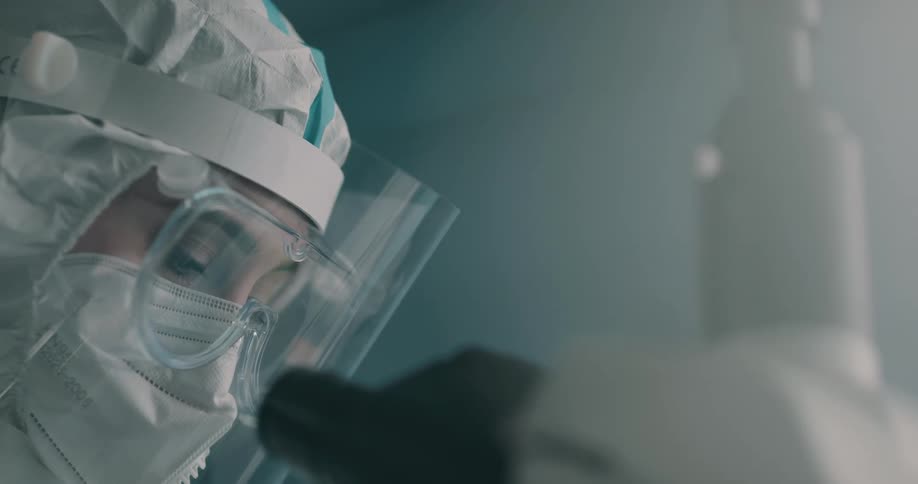 A lab worker wears a protective suit and face shield while looking through a microscope