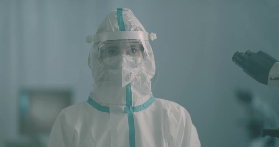 A woman with goggles and a hazmat suit on stands in front of a microscope