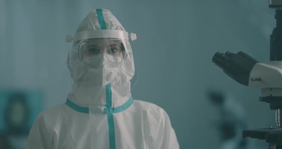 A lab worker wears a full protective suit and mask while standing near a microscope