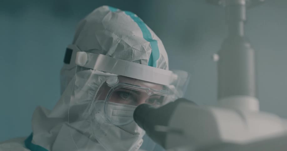 A doctor wearing a face shield over goggles and mask looks through a microscope