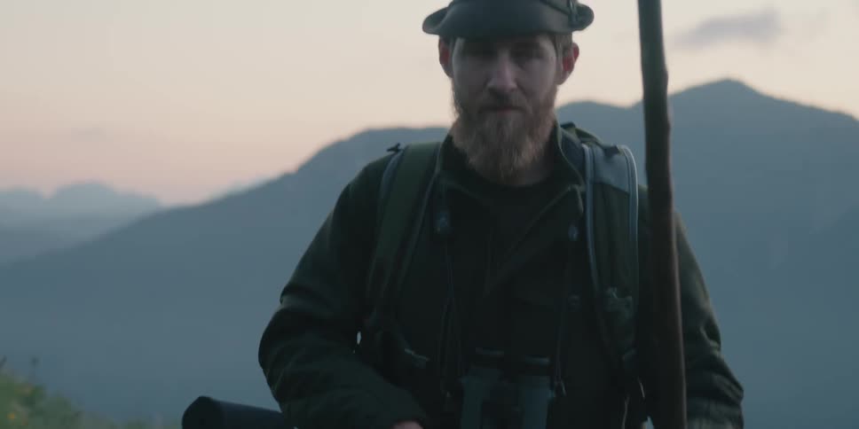 A man walks across a hill with a walking stick binoculars and a gun
