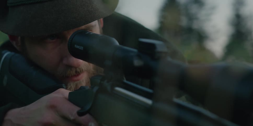 Person with beard wearing hat aims through scope on rifle