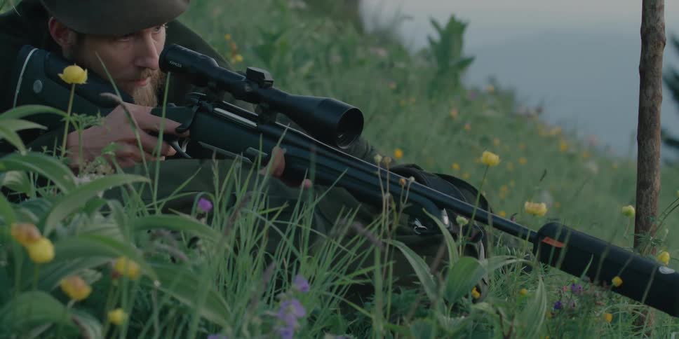 Hunter lays down over his backpack in the grass aiming a rifle with a scope