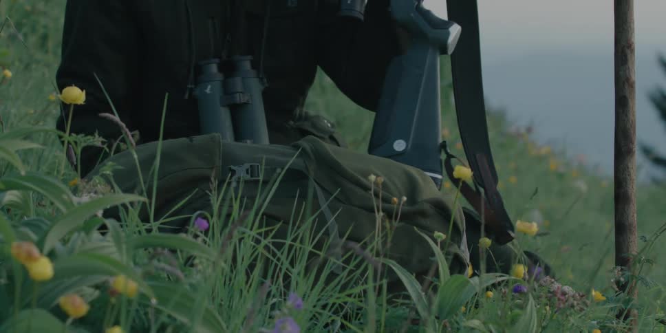 A man with a backpack and binoculars sitting down on a grassy hill and aiming a rifle
